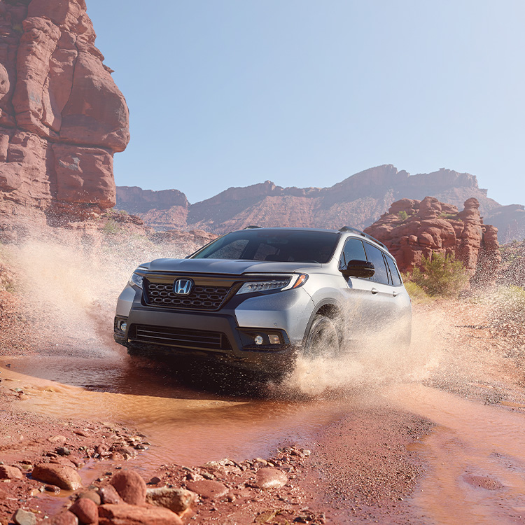 Driver-side front view of the 2021 Honda Passport Elite in Lunar Silver Metallic, demonstrating all-wheel drive and splashing through water on a rugged mountain road. 